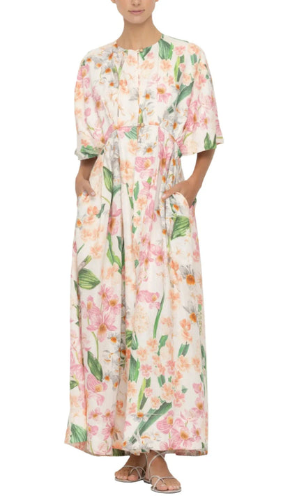 A person wearing a long, floral dress with short sleeves and sandals, standing against a plain white background showcases one of the coveted pieces: the Agua by Aguabendita Gesto Maxi Kaftan.