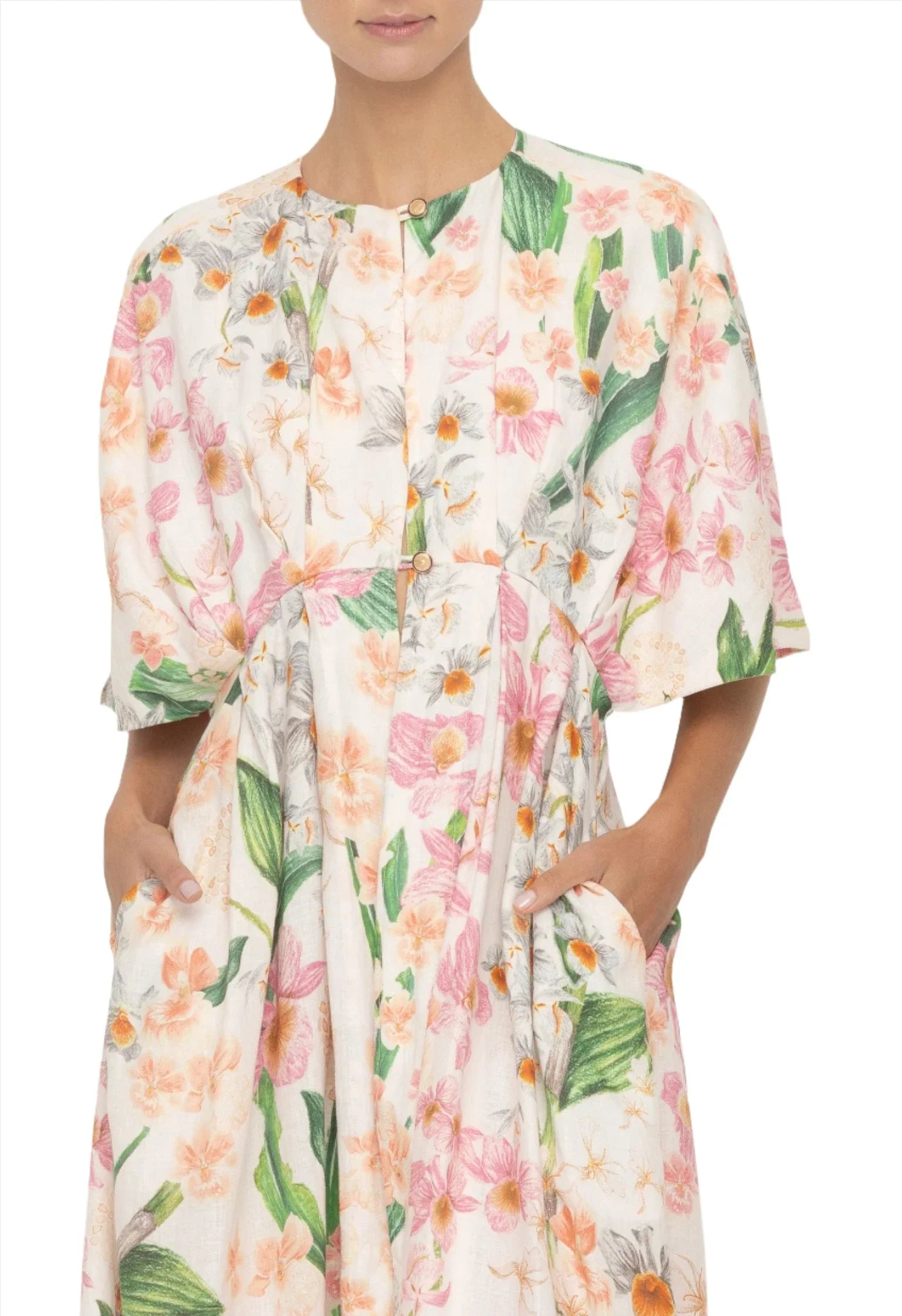 A person wearing a white floral Agua by Aguabendita Gesto Maxi Kaftan with short sleeves and front buttons. The dress, crafted from linen, features green leaves and pink to orange flowers. The person has hands in the dress pockets.