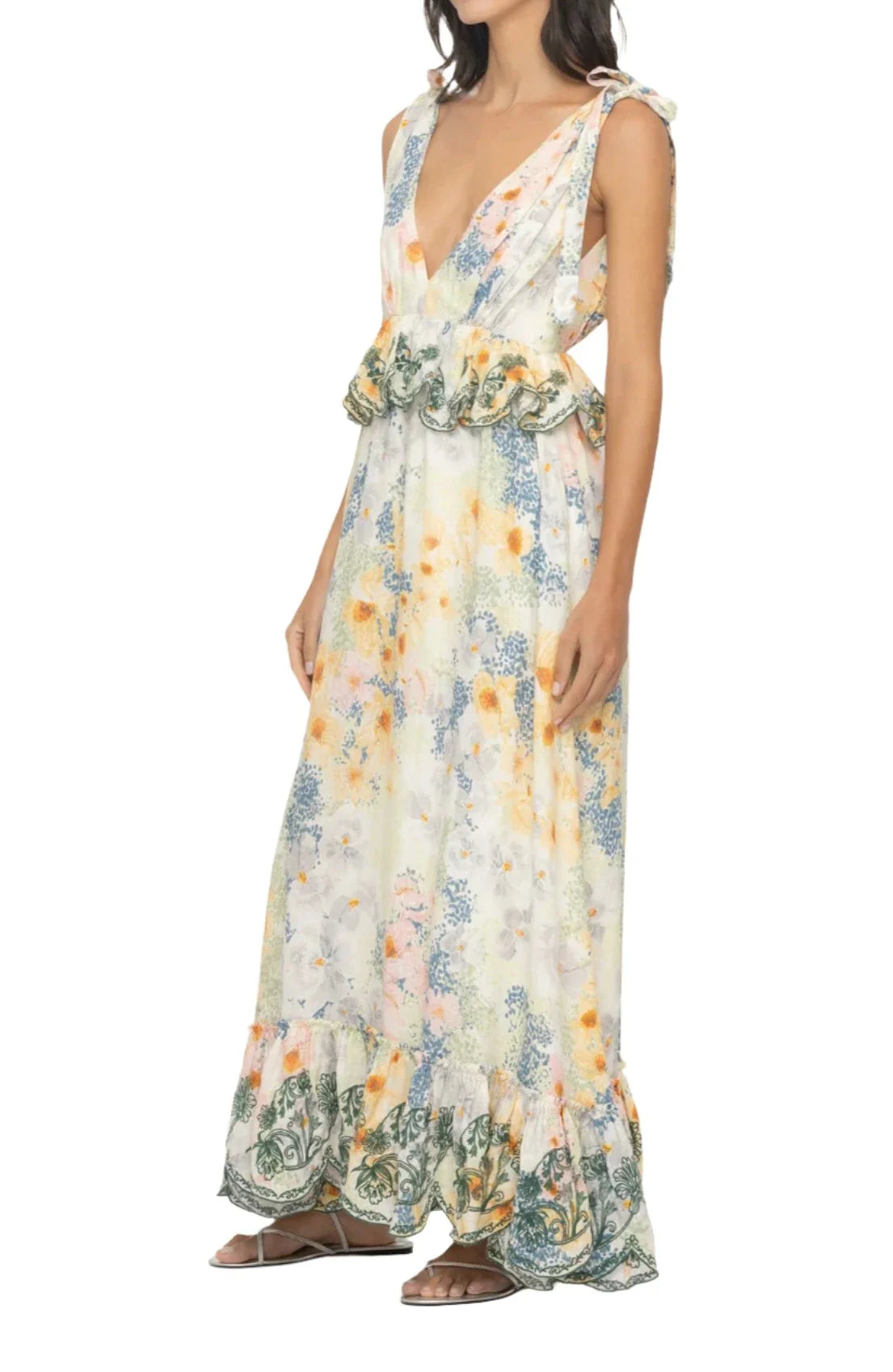 A woman wearing the Agua By Aguabendita Infinito Maxi Dress features a V-neck, floral pattern, and side cutouts. This cotton garment from Agua By Aguabendita boasts ruffled details at the waist and a ruffled hem, paired perfectly with flat sandals.