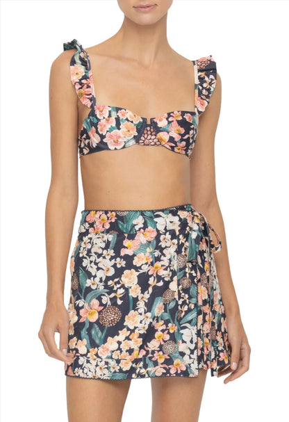 Woman wearing a floral two-piece outfit with an Agua By Aguabendita ruffled bikini top and matching Agua By Aguabendita Luna Mini Pareo skirt featuring a tie fastening.