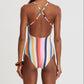 A woman stands facing away, wearing the Lemlem Retiro One Piece swimsuit with vertical multicolored stripes, adjustable cross-back straps, and a medium-coverage leg.