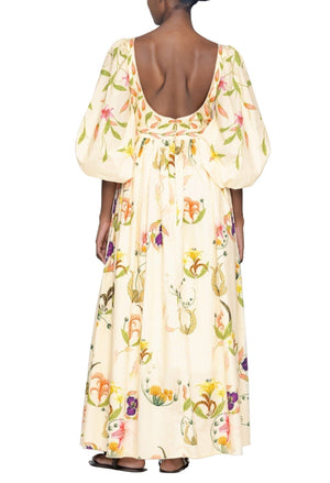 A person stands with their back to the camera wearing a long, hand-embroidered Agua by Aguabendita Vivianne Maxi Dress adorned with a floral print, featuring large puff sleeves and an open back.