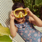 A child wearing the Louise Misha Andrea Bathing Suit, with its vintage-inspired floral design, holds a yellow snorkel mask over their face as they sit on a striped lounge chair.