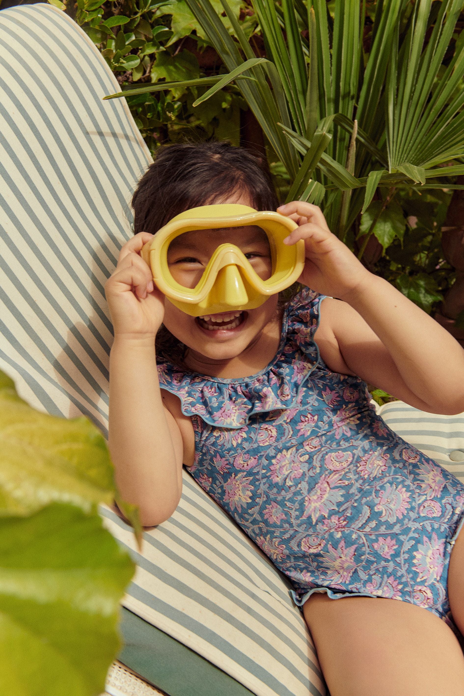 A child wearing the Louise Misha Andrea Bathing Suit, with its vintage-inspired floral design, holds a yellow snorkel mask over their face as they sit on a striped lounge chair.