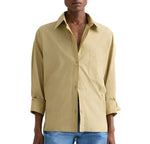 A person wearing the TWP New Earl Shirt—a relaxed, oversized light brown button-down with rolled sleeves—paired with blue jeans, stands against a plain background.