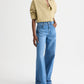 A person wears the TWP New Earl Shirt—relaxed, oversized with rolled sleeves—paired with light blue high-waisted wide-leg jeans and brown sandals, standing against a plain white background.