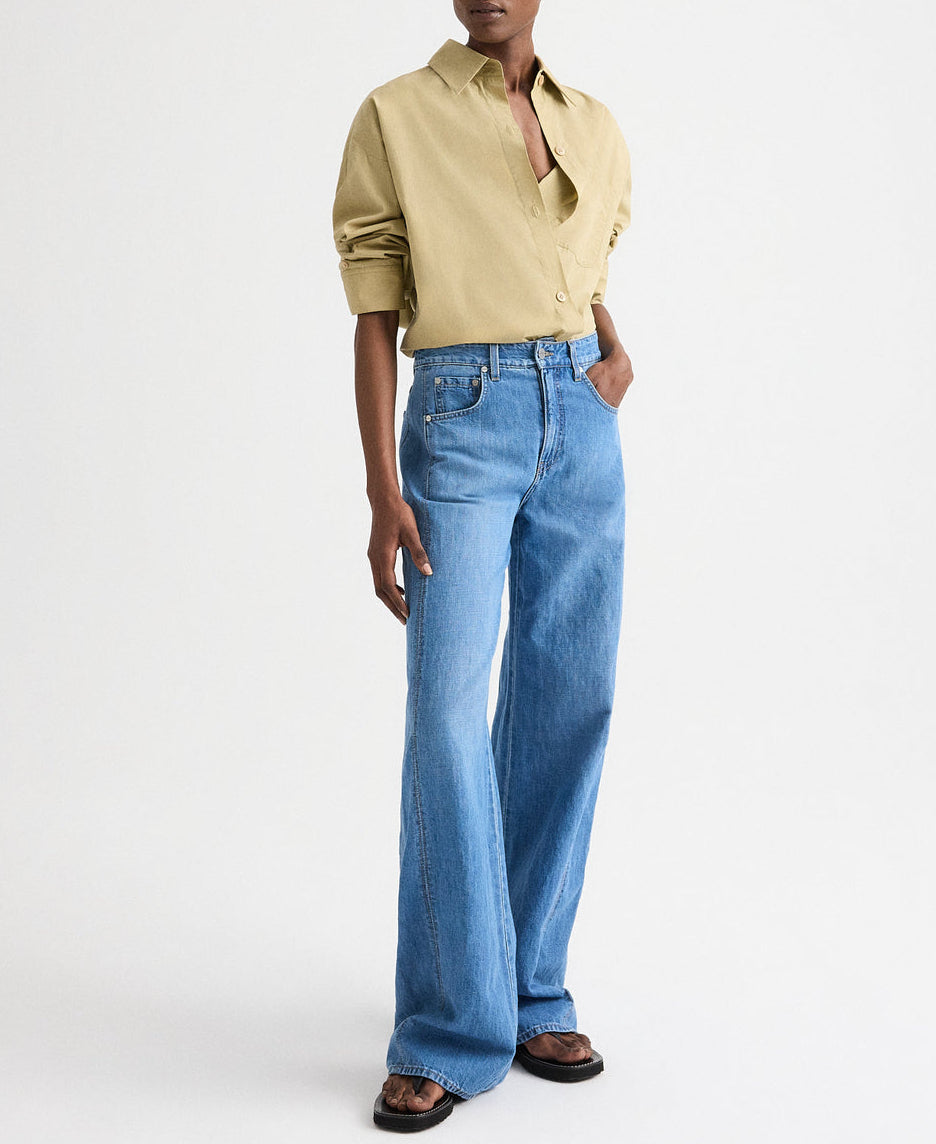 A person wears the TWP New Earl Shirt—relaxed, oversized with rolled sleeves—paired with light blue high-waisted wide-leg jeans and brown sandals, standing against a plain white background.