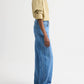 A person stands sideways against a plain white background, wearing the TWP New Earl Shirt—relaxed striped Italian cotton with rolled sleeves—paired with blue wide-leg jeans and black sandals.