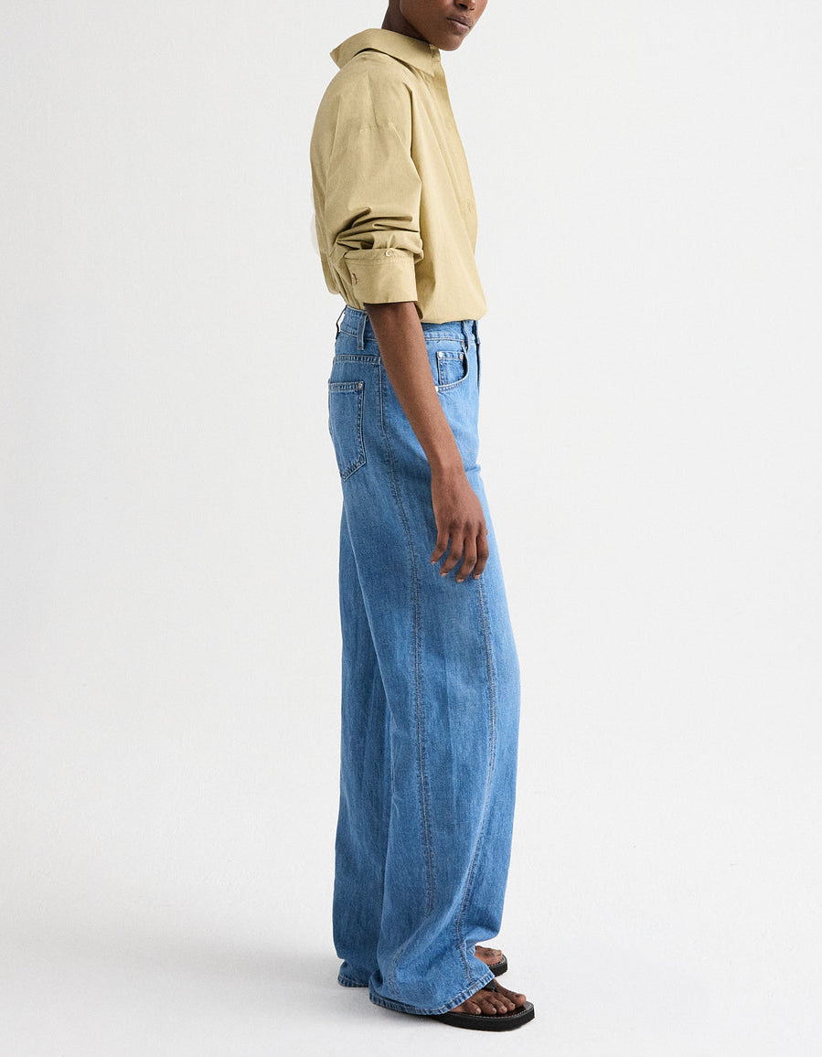 A person stands sideways against a plain white background, wearing the TWP New Earl Shirt—relaxed striped Italian cotton with rolled sleeves—paired with blue wide-leg jeans and black sandals.