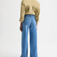 A person stands facing away, wearing the TWP New Earl Shirt in light brown and high-waisted, wide-leg blue jeans against a plain light background.