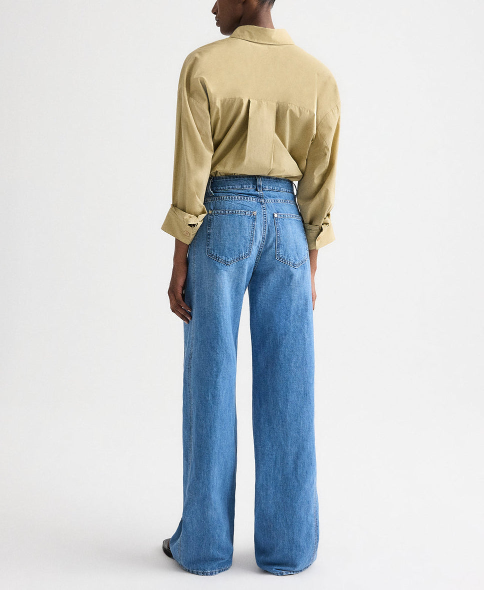 A person stands facing away, wearing the TWP New Earl Shirt in light brown and high-waisted, wide-leg blue jeans against a plain light background.