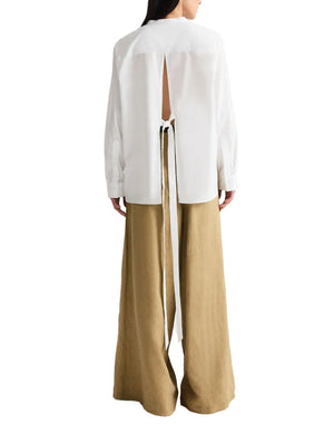 A model wears the TWP Tied Down Shirt, a white long-sleeve top with a back slit and tie detail inspired by classic cotton shirting, paired with wide-leg tan pants against a white background.