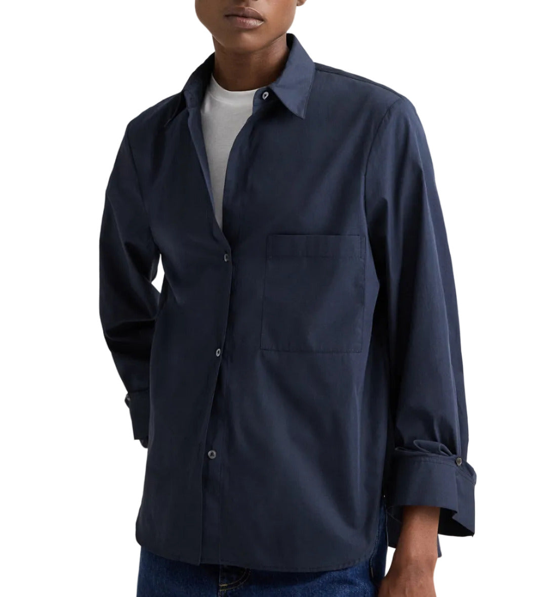 A person wearing the TWP Next Morning Shirt, an oversized navy blue Italian cotton button-down, layered over a white t-shirt with blue jeans and standing against a plain background.