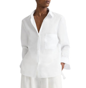 A person wears the TWP Next Morning Shirt, an oversized Italian cotton button down with a relaxed fit and single chest pocket, paired with white pants and shown from mid-thigh up.