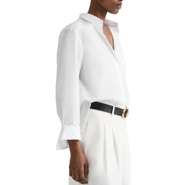 A person wears the TWP Next Morning Shirt, an oversized Italian cotton button-down, tucked into high-waisted white trousers with a black belt and round gold buckle for a refined yet relaxed look.