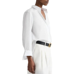 A person wears the TWP Next Morning Shirt, an oversized Italian cotton button-down, tucked into high-waisted white trousers with a black belt and round gold buckle for a refined yet relaxed look.