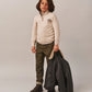 A young boy wears the Bonpoint Boys' Gert Cashmere Sweater with a trucker collar and Bonpoint embroidery, paired with plaid shirt, olive pants, and brown boots, standing against a plain light background holding a dark jacket.