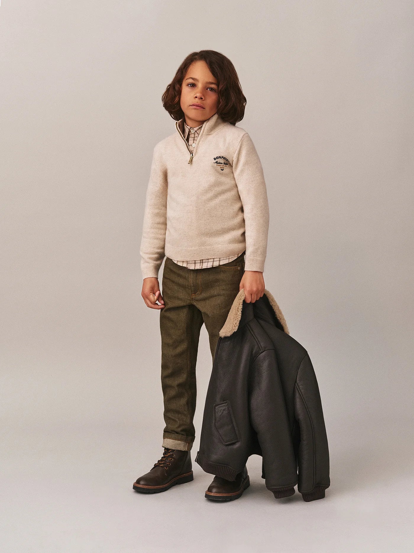A young boy wears the Bonpoint Boys' Gert Cashmere Sweater with a trucker collar and Bonpoint embroidery, paired with plaid shirt, olive pants, and brown boots, standing against a plain light background holding a dark jacket.