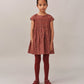 A young girl faces forward in a Bonpoint Girls' Duchesse Smocked Dress made with Liberty Fabric, paired with matching red tights and shoes, set against a plain light background.