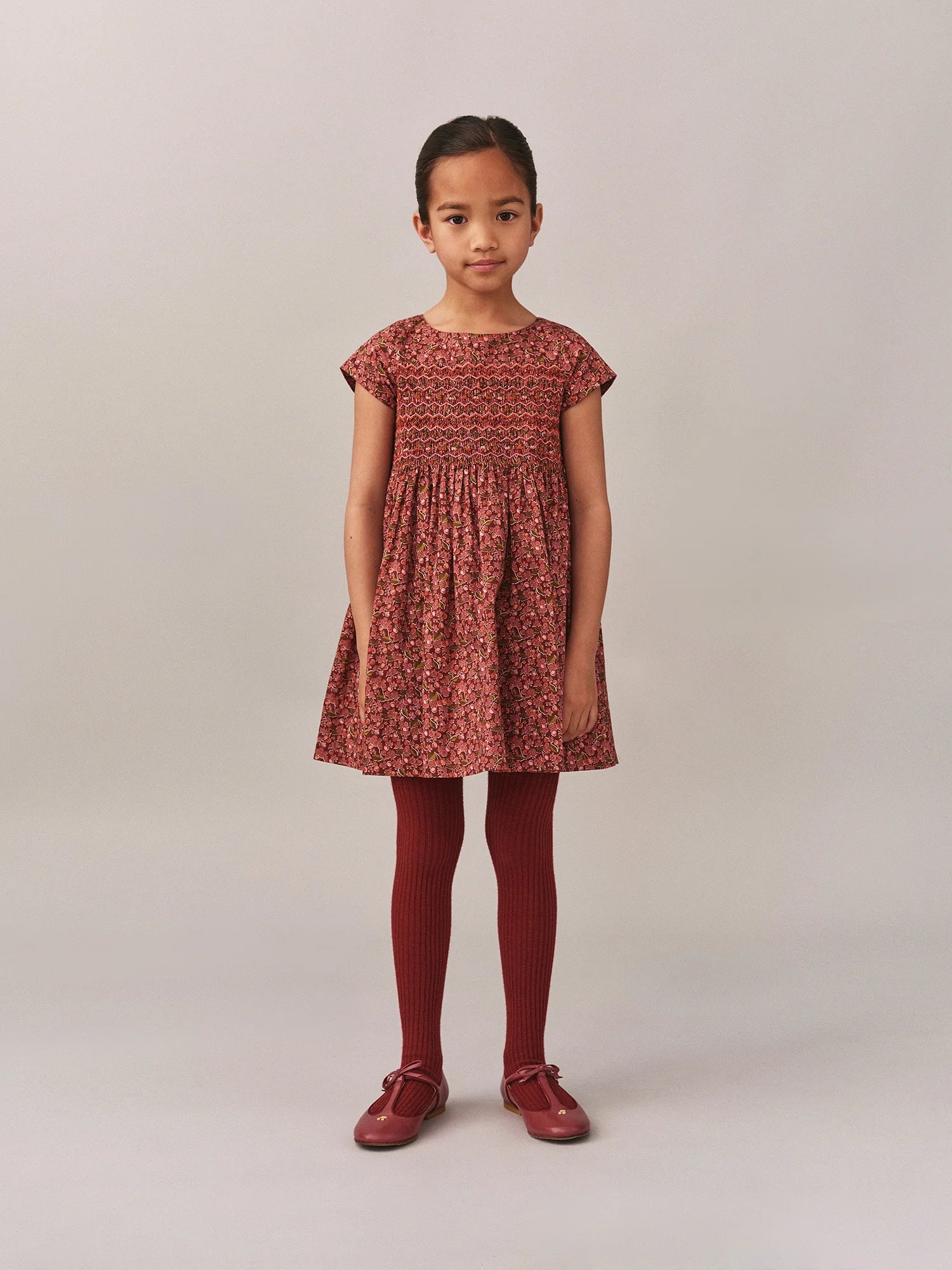 A young girl faces forward in a Bonpoint Girls' Duchesse Smocked Dress made with Liberty Fabric, paired with matching red tights and shoes, set against a plain light background.