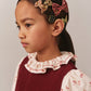 A young girl in a maroon dress with a ruffled floral collar wears multiple girls' hair accessories, including the Bonpoint Tutu Hair Clip made with Liberty Fabric, as she looks to the side against a plain background.