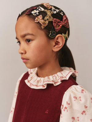 A young girl in a maroon dress with a ruffled floral collar wears multiple girls' hair accessories, including the Bonpoint Tutu Hair Clip made with Liberty Fabric, as she looks to the side against a plain background.