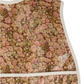 Close-up of the Bonpoint Jlip Adjustable Bib, crafted with Liberty Fabrics, showcasing a brown, pink, and cream floral pattern with white trim on the edges and front pocket.