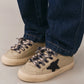 A person wearing dark blue jeans and Bonpoint x Golden Goose baby sneakers, featuring beige uppers, black star patches, and black-and-white laces, stands on a light surface.