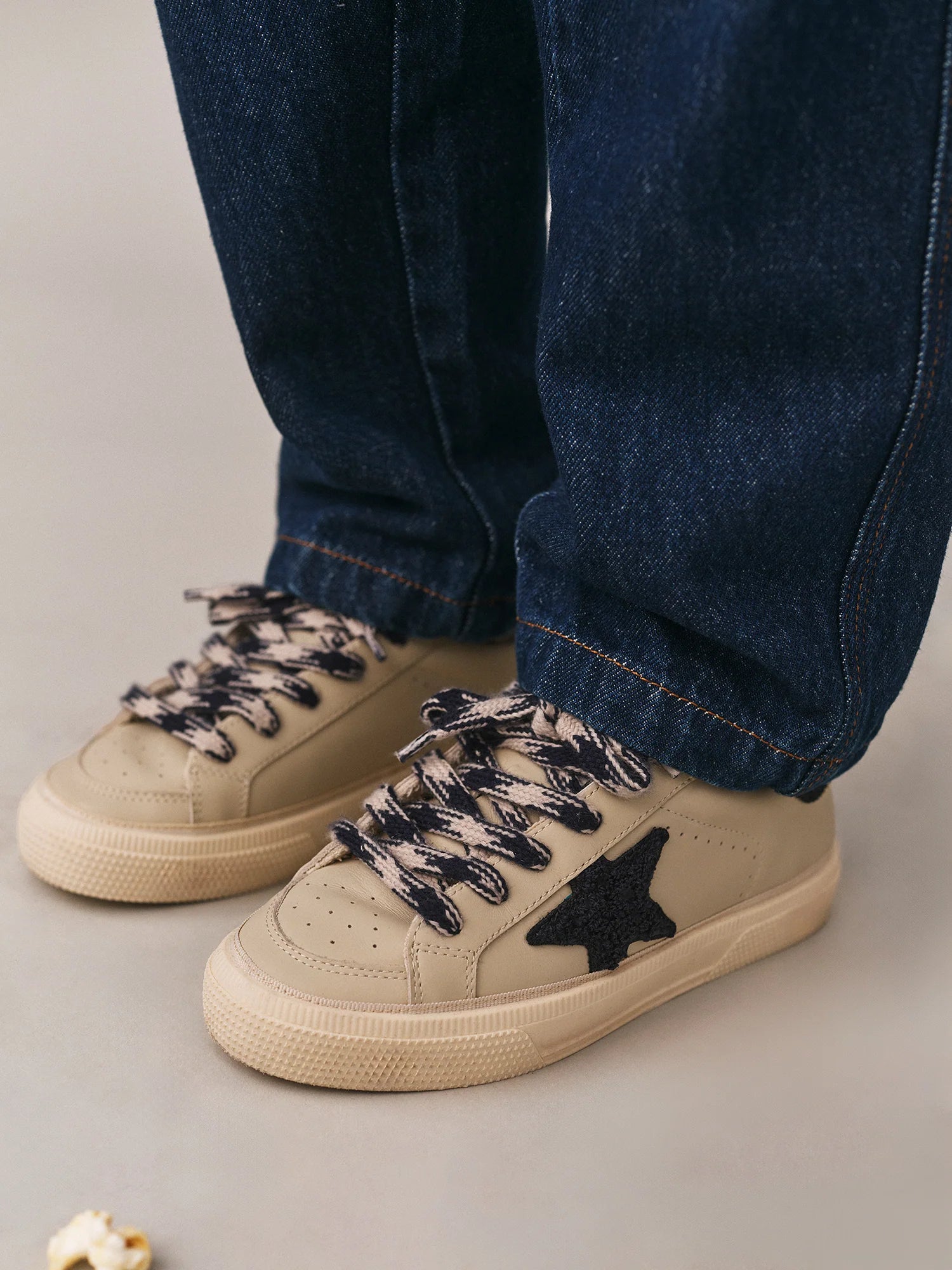 A person wearing dark blue jeans and Bonpoint x Golden Goose baby sneakers, featuring beige uppers, black star patches, and black-and-white laces, stands on a light surface.