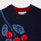 The Girls’ Maxine Sweater by Bonpoint is a navy blue knit girls sweater featuring a red and blue intarsia-knit cherries and leaf pattern near the collar, a ribbed neckline, and an exposed Maxine sweater label.