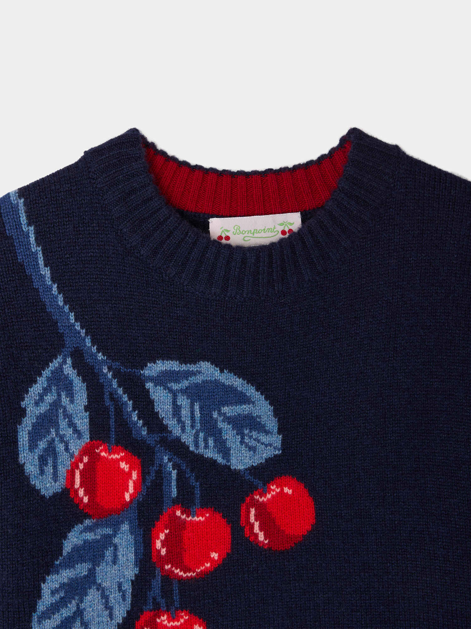 The Girls’ Maxine Sweater by Bonpoint is a navy blue knit girls sweater featuring a red and blue intarsia-knit cherries and leaf pattern near the collar, a ribbed neckline, and an exposed Maxine sweater label.