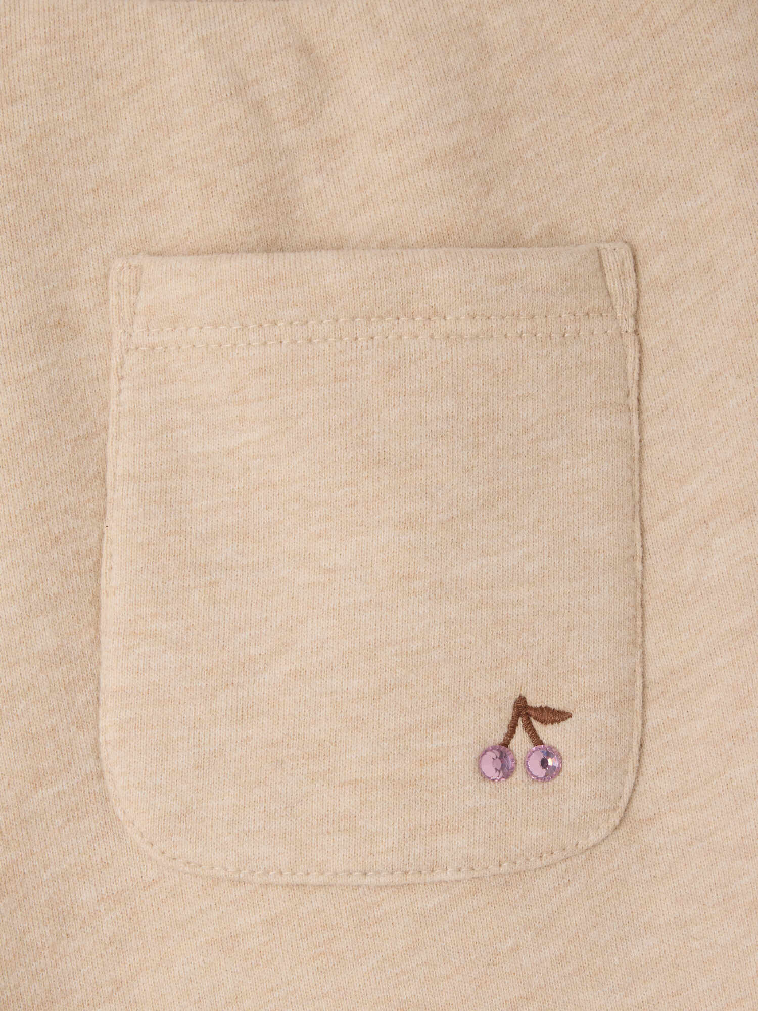Close-up of a beige fabric pocket on the Bonpoint Girls’ Dalila Joggers, featuring a small embroidered cherry design with two pink sequins as the fruit. These organic cotton joggers combine comfort with playful detail.