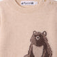 The Bonpoint Baby and Kids’ Almire Pullover is a beige knit sweater with two shoulder buttons, featuring a charming graphic of a seated brown bear on the front.