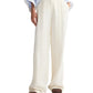 A person wearing TWP's Hudson St Pant—relaxed mid-rise, wide-leg white trousers—paired with a light blue rolled-cuff shirt and brown shoes, stands hands in pockets.