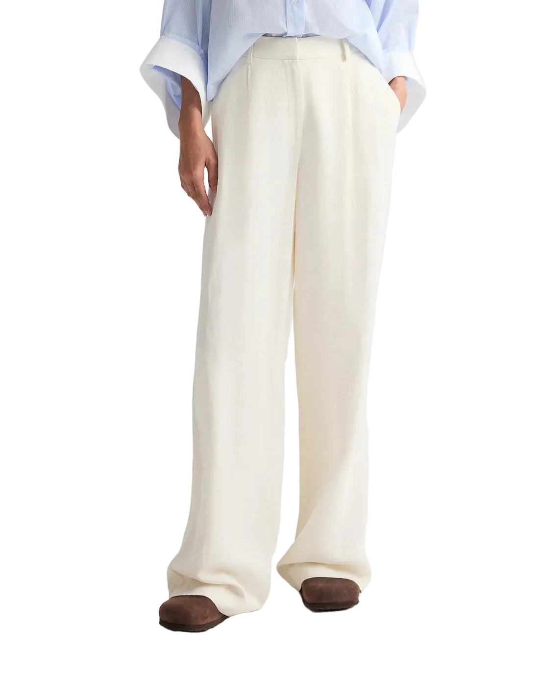 A person wearing TWP's Hudson St Pant—relaxed mid-rise, wide-leg white trousers—paired with a light blue rolled-cuff shirt and brown shoes, stands hands in pockets.