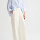 A person stands against a plain background, hands in pockets, wearing the TWP Hudson St Pant—relaxed mid-rise, wide-leg pants in coated viscose linen—with a light blue oversized shirt.