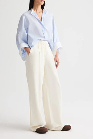 A person stands against a plain background, hands in pockets, wearing the TWP Hudson St Pant—relaxed mid-rise, wide-leg pants in coated viscose linen—with a light blue oversized shirt.