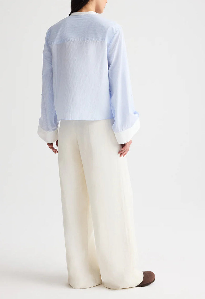 A person stands facing away, wearing TWP’s Hudson St Pant—relaxed mid-rise, wide-leg pants in a coated viscose linen blend—paired with a light blue long-sleeve shirt with white cuffs, set against a plain light background.