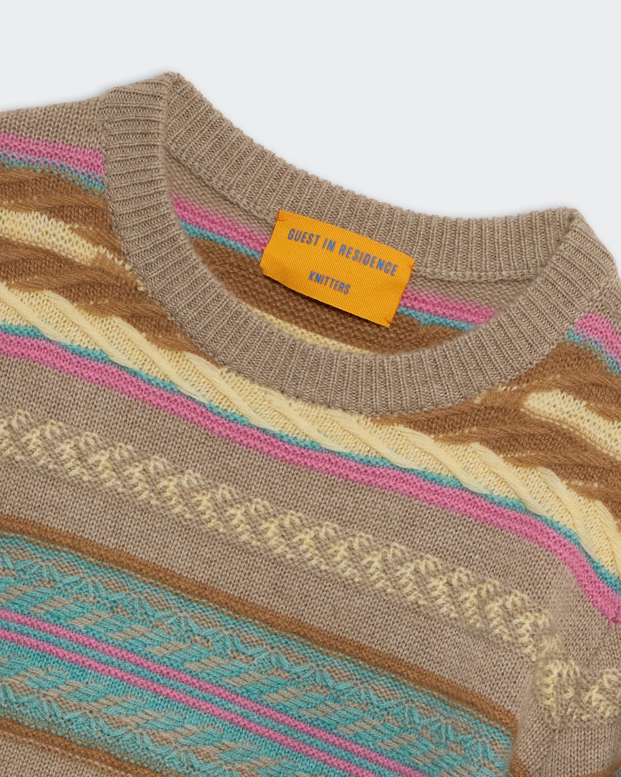 Close-up of the Western Stripe Crew sweater by Guest in Residence, featuring multicolored jacquard stitch, cable patterns, and a yellow "Guest in Residence Knitters" tag on the neckline.
