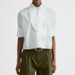 Person wearing the TWP Tie One On Shirt in white Supima cotton with rolled-up sleeves and oversized collar, paired with olive green high-waisted pleated pants, a black belt, and gold buckle.