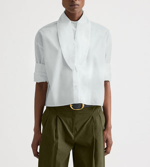 Person wearing the TWP Tie One On Shirt in white Supima cotton with rolled-up sleeves and oversized collar, paired with olive green high-waisted pleated pants, a black belt, and gold buckle.