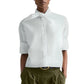 A person wears the TWP Tie One On Shirt—a vintage washed poplin white button-up with a tied neck detail and rolled-up sleeves—paired with olive green pleated pants and a black belt.