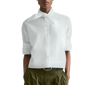 A person wears the TWP Tie One On Shirt—a vintage washed poplin white button-up with a tied neck detail and rolled-up sleeves—paired with olive green pleated pants and a black belt.