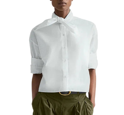 A person wears the TWP Tie One On Shirt—a vintage washed poplin white button-up with a tied neck detail and rolled-up sleeves—paired with olive green pleated pants and a black belt.