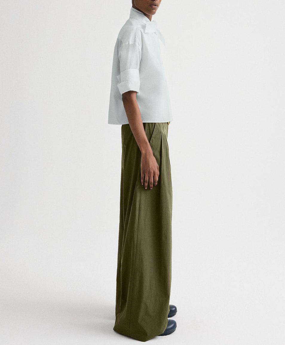 Person standing sideways wearing a boxy white Tie One On Shirt by TWP, wide olive-green pants, and black shoes against a plain light background.
