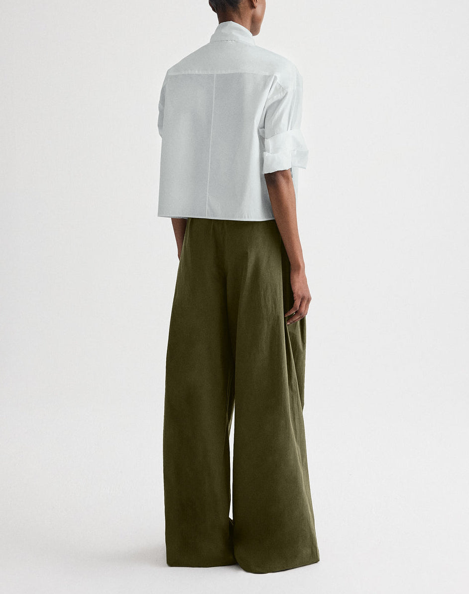 Person standing against a white background, wearing the TWP Tie One On Shirt—a light-colored cropped button down with rolled sleeves—and wide-leg olive green pants made from vintage washed poplin.