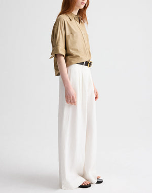 A person wears the TWP Avery Pant, styled with a loose tan button-up shirt, black belt, and black sandals, standing against a plain light background.