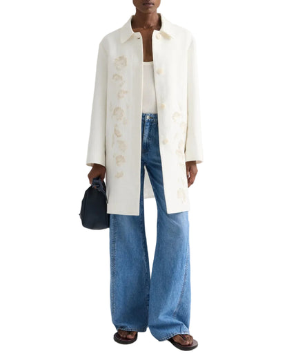 A person wearing the TWP Edinburgh Coat over a white top, with wide-leg blue jeans, brown sandals, and holding a small dark handbag.