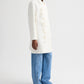 A person wears the TWP Edinburgh Coat with light floral embroidery, paired with blue wide-leg jeans and brown sandals, standing against a plain white background.
