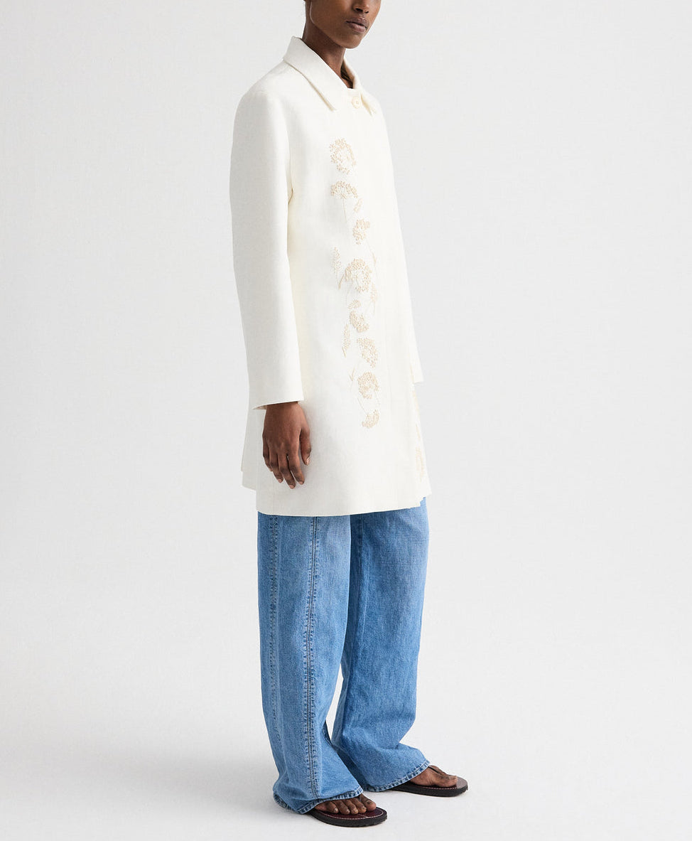 A person wears the TWP Edinburgh Coat with light floral embroidery, paired with blue wide-leg jeans and brown sandals, standing against a plain white background.